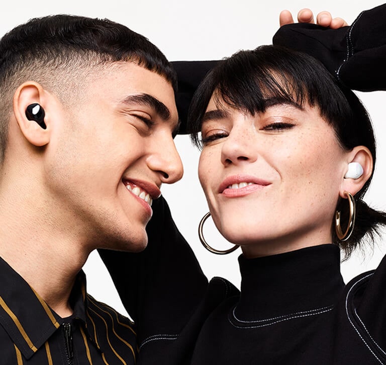 A man wearing black Galaxy Buds plus smiles as he looks a woman next to him wearing white Galaxy Buds plus. The woman smiles with her hands in the air behind her head.