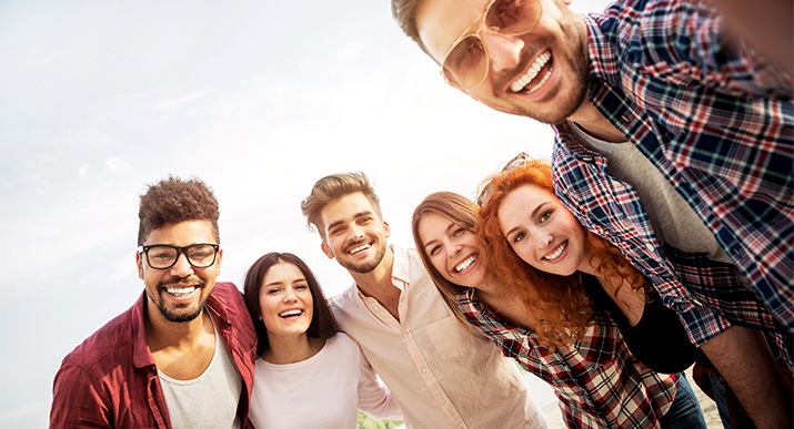 Un grupo de tres hombres y tres mujeres jóvenes están abrazados mirando hacia el frente para tomarse una selfie grupal.
