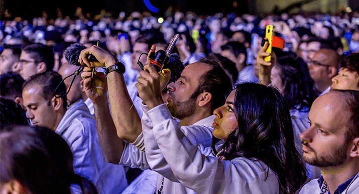 In the midst of a crowd attending an event, a man and a woman are recording the event with their smartphones that have mic attachments.