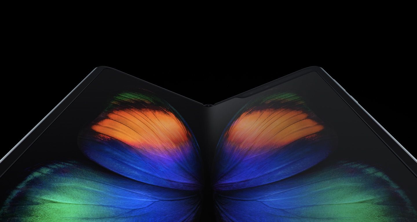 Galaxy Fold, unfolded and laying at a slight angle seen from the bottom with a green, orange, and blue butterfly graphic on-screen. It starts with an up-close shot on the Infinity Flex Display and zooms out to show the whole phone