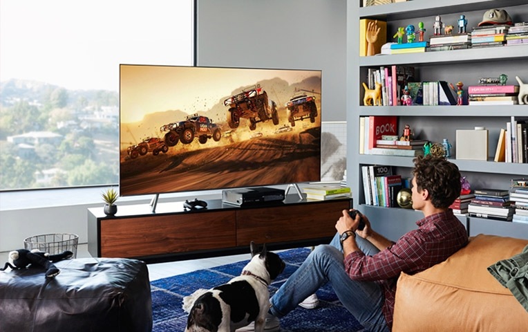 A lifestyle image of the 2018 new QLED TV Q6F. QLED TV is put on a table in the living room and a young man is playing game with QLED TV.