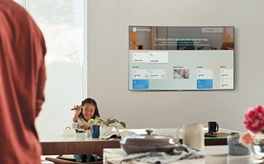 A lifestyle image of the 2018 new QLED TV Q8C. Image shows QLED TV is mounted on wall with IoT Dashboard on. A Woman is managing IoT Dashboard from the kitchen and a child is sitting in front of the TV.