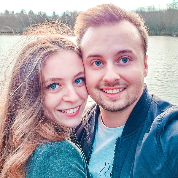 Un couple d’amoureux aux yeux bleus sourit tout en prenant un selfie devant une étendue d’eau