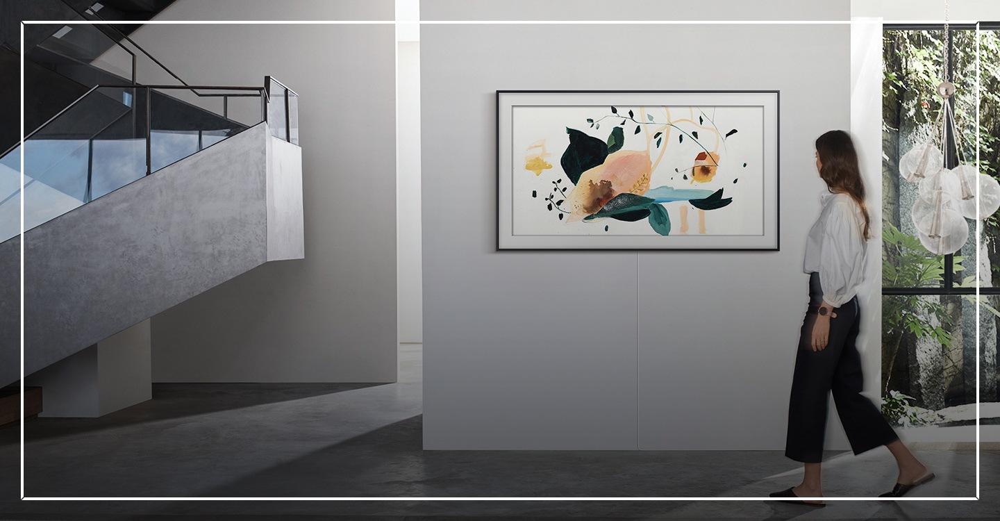 A woman walks by The 2022 Samsung The Frame TV mounted on the wall, looking like art as it displays a painting in Art Mode.