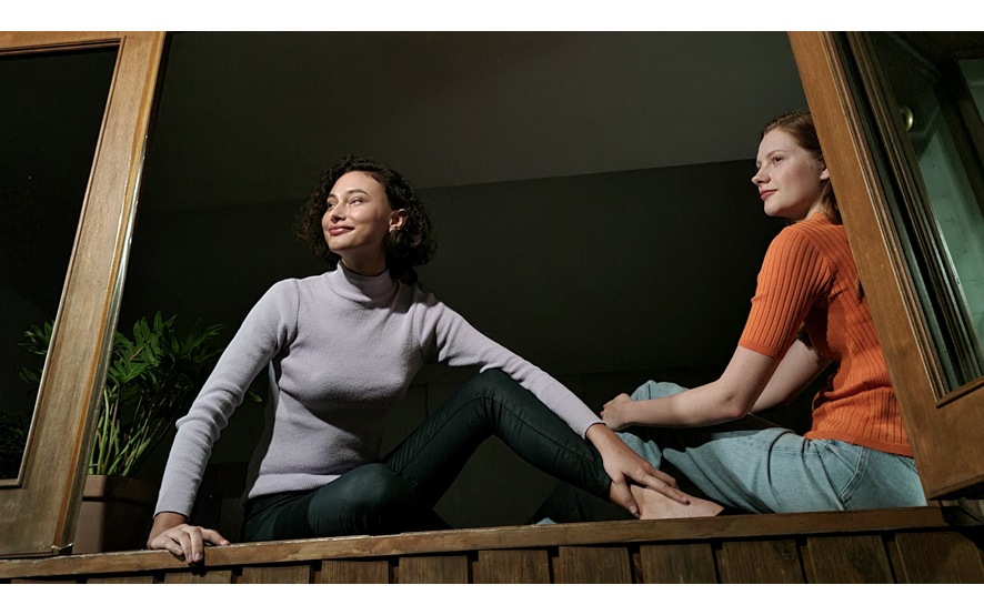 Two women sitting on a wooden ledge of an open window. The woman on the left is wearing a lavender turtleneck shirt with dark jeans and the woman on the right is wearing an orange t-shirt with light blue jeans. Their gaze is focused to the left.