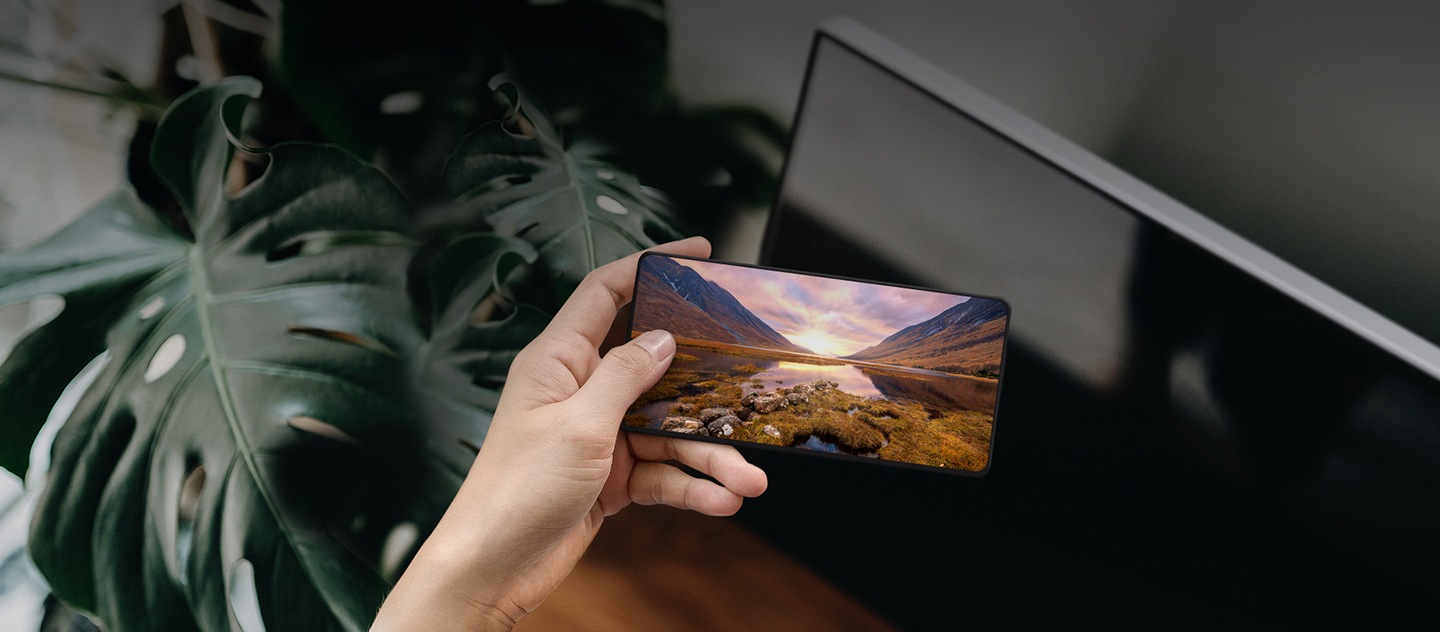 Uma mão a segurar um smartphone Galaxy na frente de uma TV Samsung. O ecrã do Galaxy mostra uma imagem de uma paisagem majestosa. 