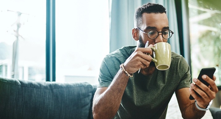 Um homem está sentado num sofá a tomar um café, enquanto observa o smartphone na sua mão esquerda.