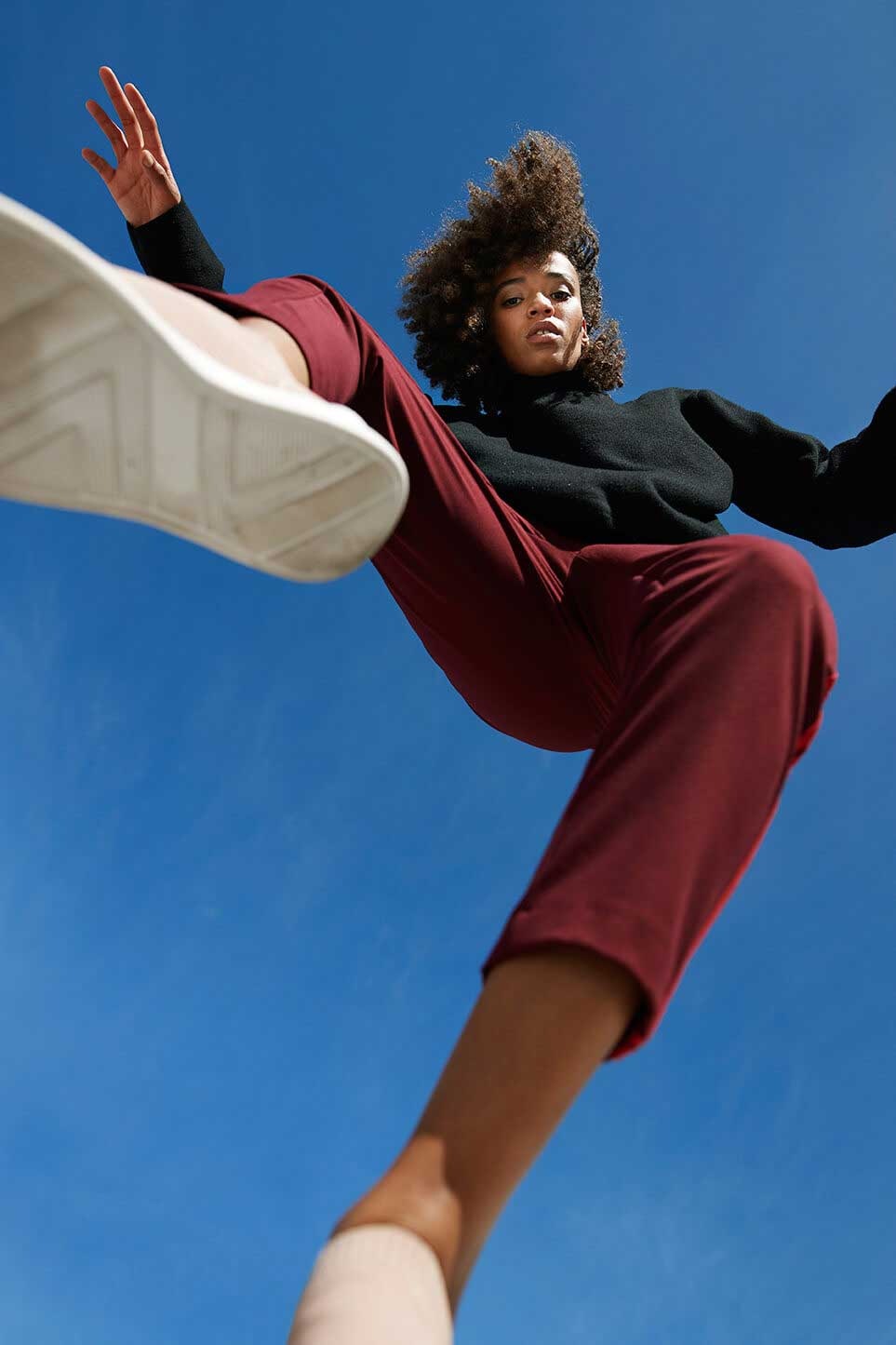 Uma mulher faz uma pose enquanto dança e olha para a câmara em baixo. Ao fundo está um céu azul limpo.