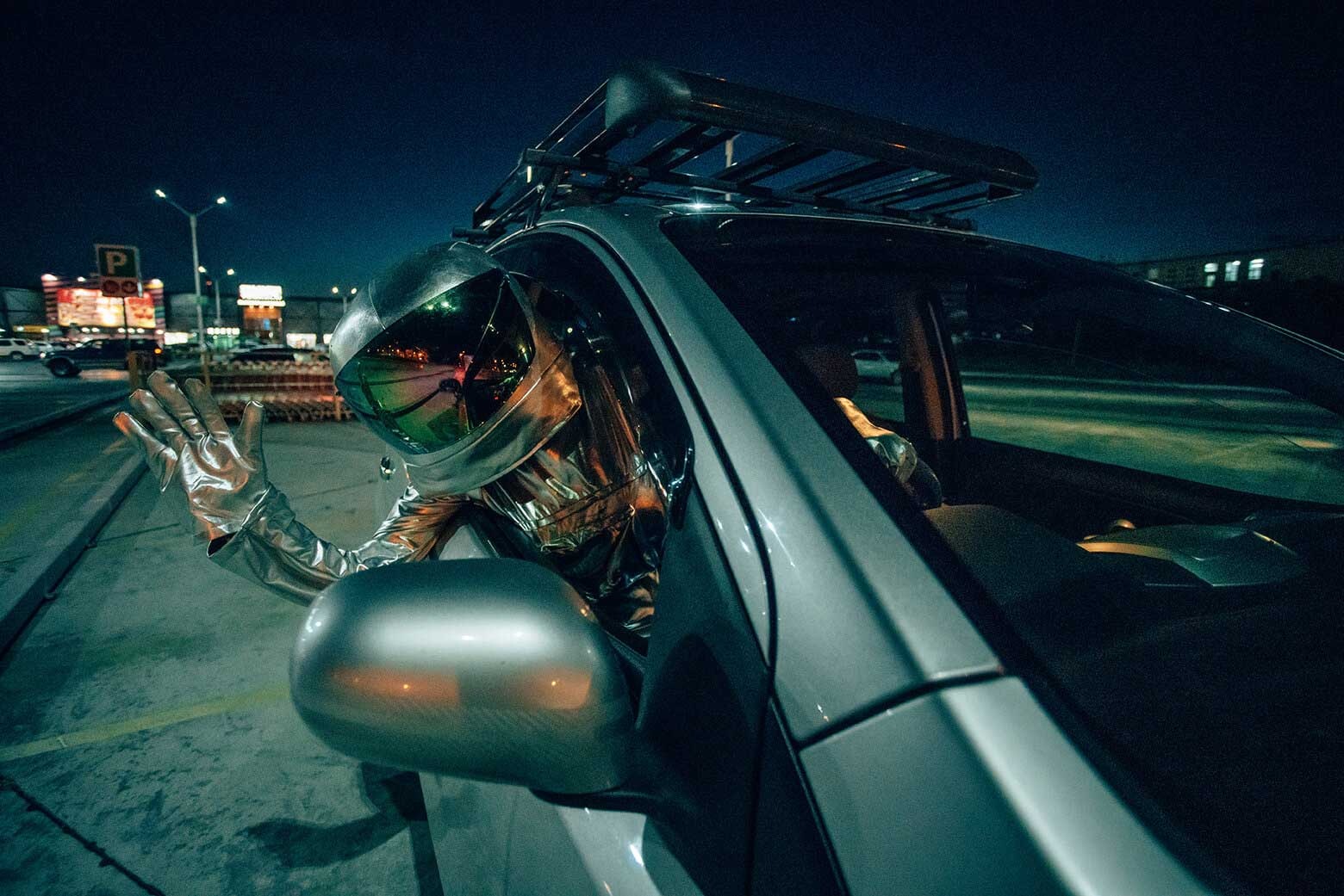 Uma pessoa vestida de astronauta espreita pela janela de um carro prateado e acena.