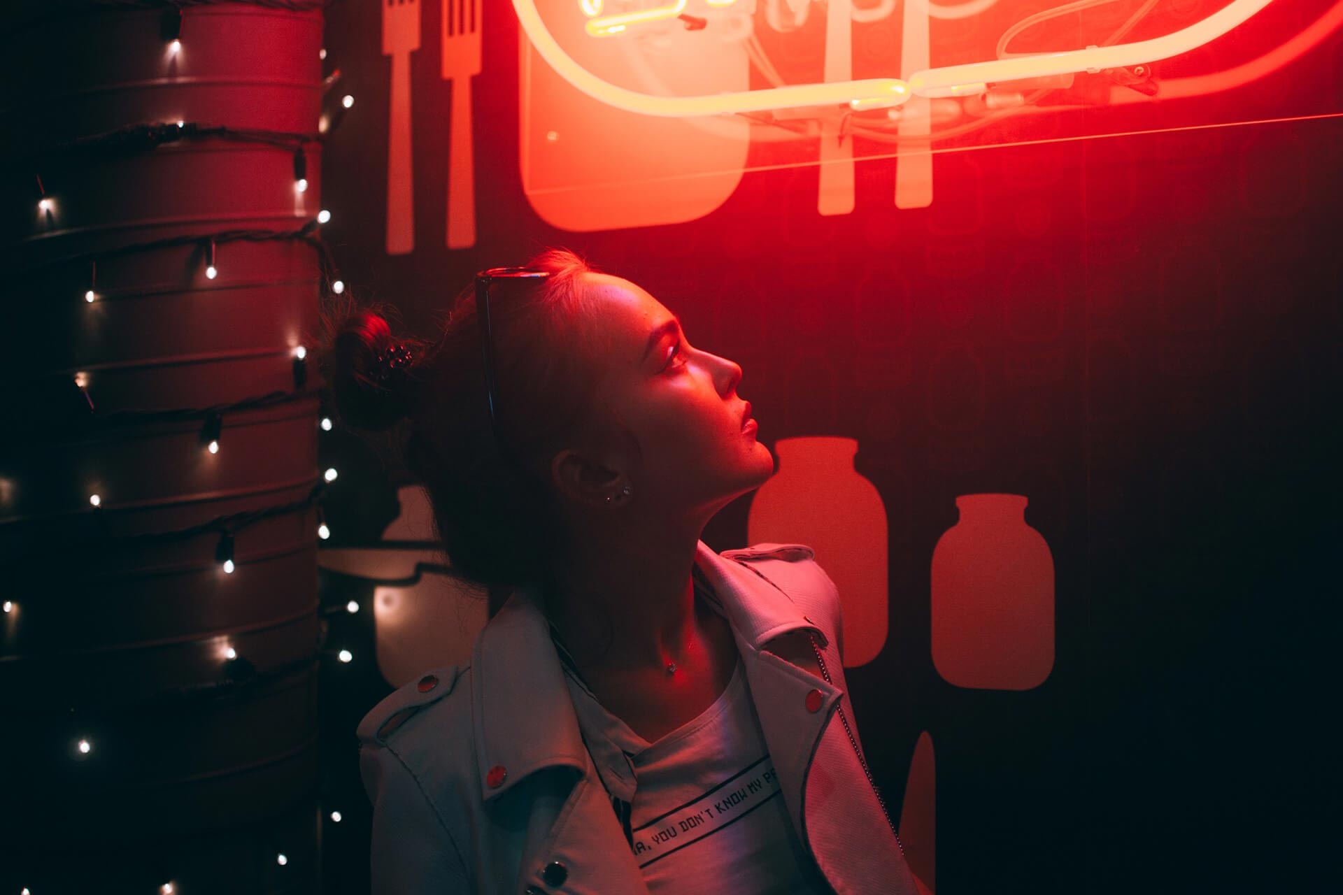 Uma mulher olha para a esquerda para uma janela que é iluminada por um sinal néon vermelho brilhante. Existem luzes etéreas à sua esquerda.