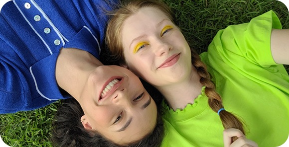 An aerial view of two women laying side-by-side on the grass. The woman on the left is wearing a blue collared shirt while the woman on the right is wearing a lime green top. Their heads are pressed together from opposite ends. They're both smiling at the camera