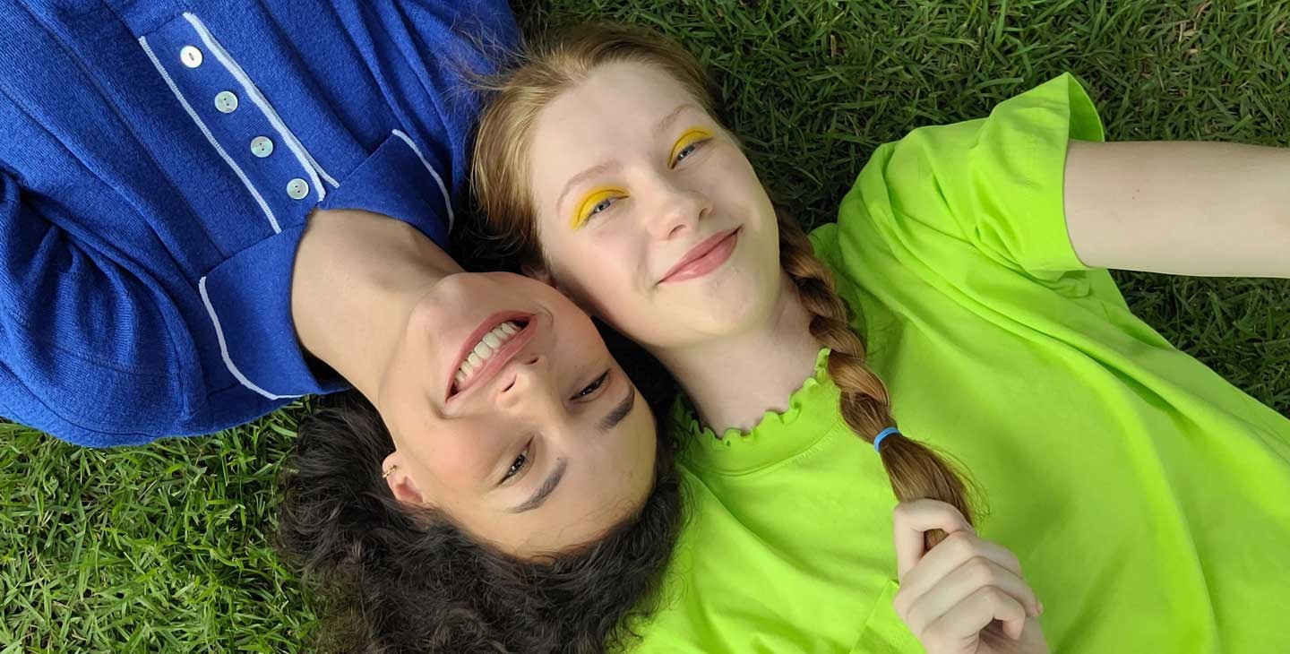 An aerial view of two women laying side-by-side on the grass. The woman on the left is wearing a blue collared shirt while the woman on the right is wearing a lime green top. Their heads are pressed together from opposite ends. They're both smiling at the camera.