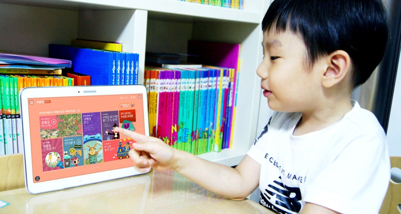 한 어린이가 책상에 앉아 웅진북클럽 화면이 나오는 태블릿을 손으로 터치하는 모습을 보여주고 있습니다.