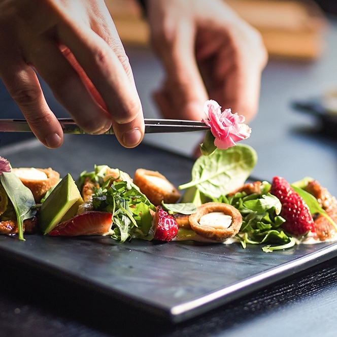 검정색 플레이팅 도마 위에 샐러드가 놓여 있고, 핀셋으로 꽃을 들고 샐러드에 장식하려고 하는 모습입니다.