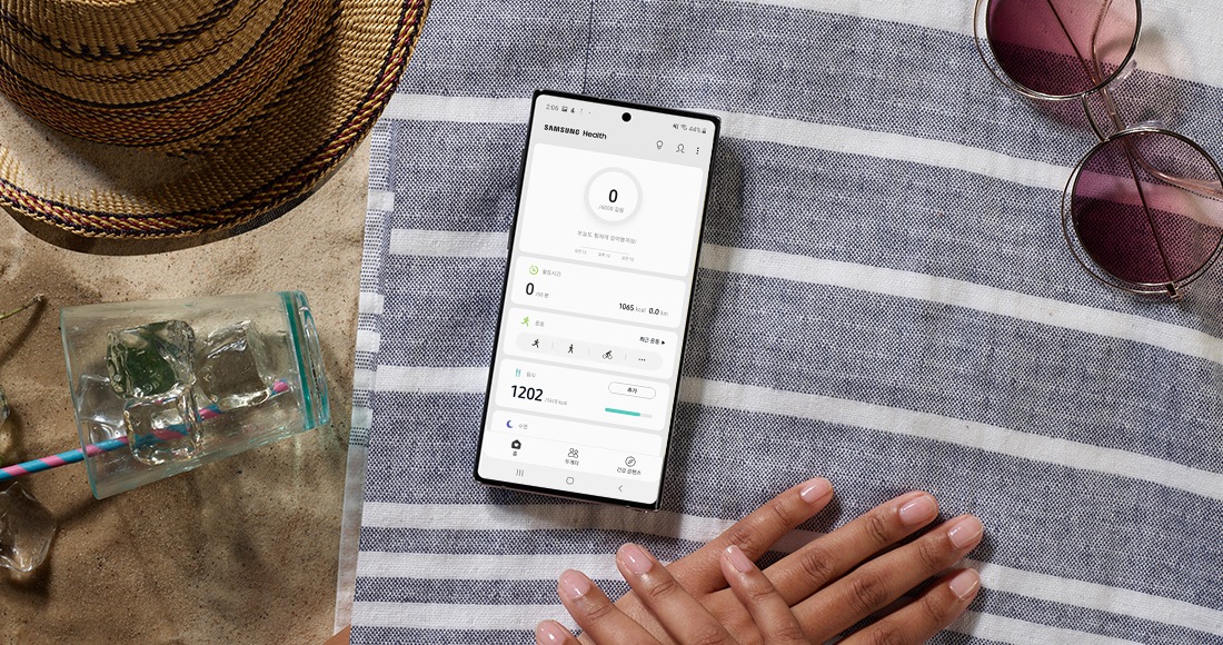 Samsung Health가 실행된 갤럭시 노트10+가 해변에 깔려진 매트 위에 놓여져 있습니다. 배경에는 모자, 선글라스, 얼음이 담긴 컵이 보이며 휴가 때도 건강 데이터를 추적하는게 쉽다는 것을 보여줍니다.