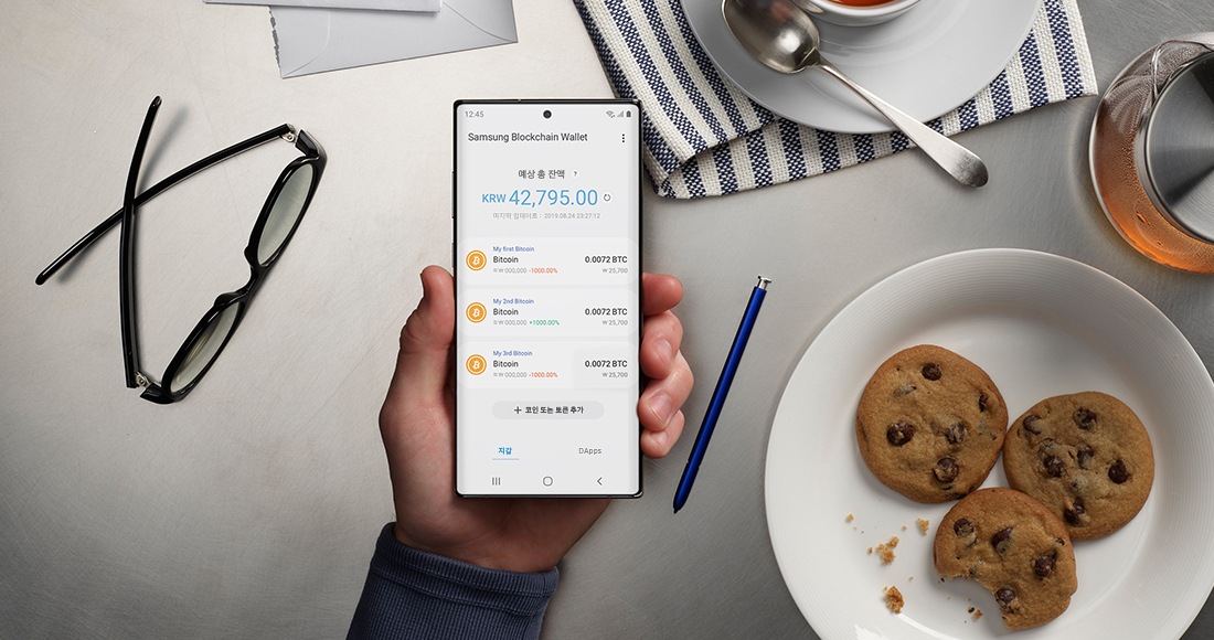 Samsung Blockchain Wallet이 실행된 갤럭시 노트10+를 손에 들고 있습니다. 배경에는 안경, 과자가 담긴 접시, 차 등이 있으며 집에서도 블록체인 거래를 쉽고 안전하게 체결할 수 있는 모습을 보여줍니다.