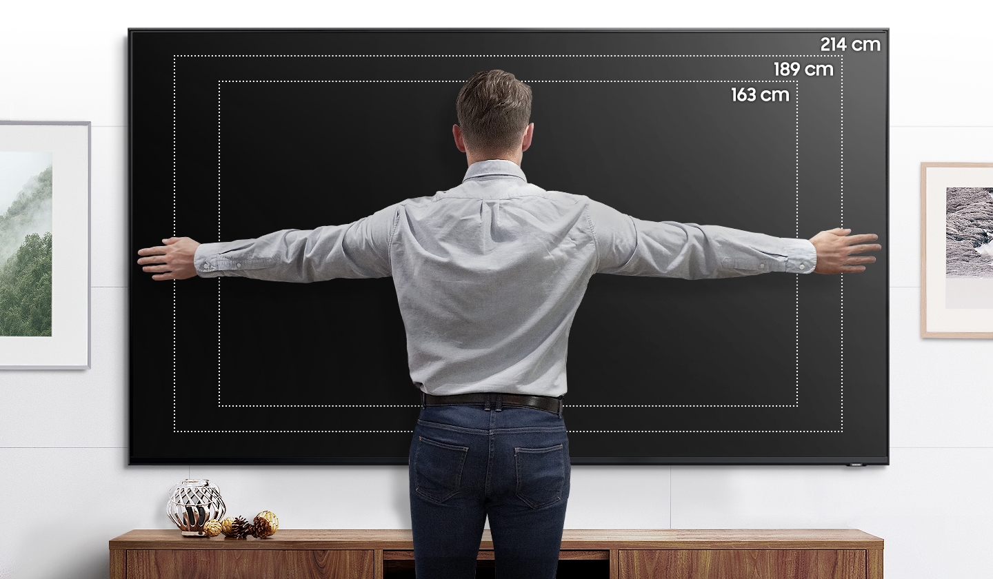한 남자가 TV가 크다는 것을 보여주기 위해 팔을 벌린 채 75인치 TV에 장착된 벽 앞에 서 있습니다.
