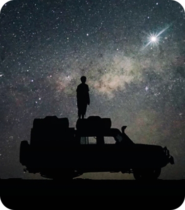 별빛이 장관을 이루는 밤 하늘을 배경으로 한 사람이 차 위에 서서 하늘을 바라보고 있습니다.