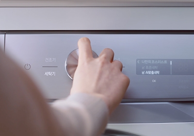 세탁기의 조작 패널을 돌리고 있는 여성의 손을 클로즈업했습니다. 조작 패널은 AI 맞춤 세탁을 실행하고 있다는 것을 설명합니다.