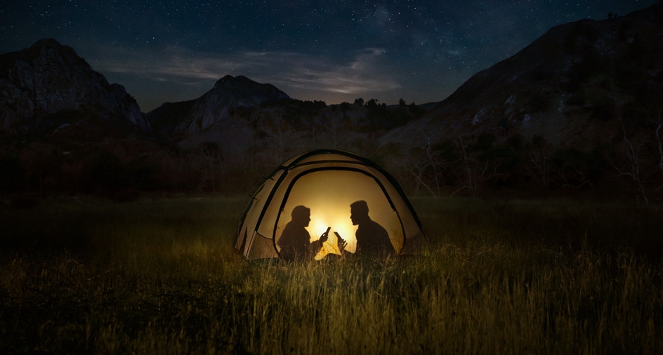 어두운 산 속의 텐트 안에 두 명의 사람이 있습니다. 텐트 안엔 불빛이 밝혀져 있고, 두 사람은 스마트폰을 들고 있습니다. 배터리가 오래 간다는 것이 잘 드러납니다.
