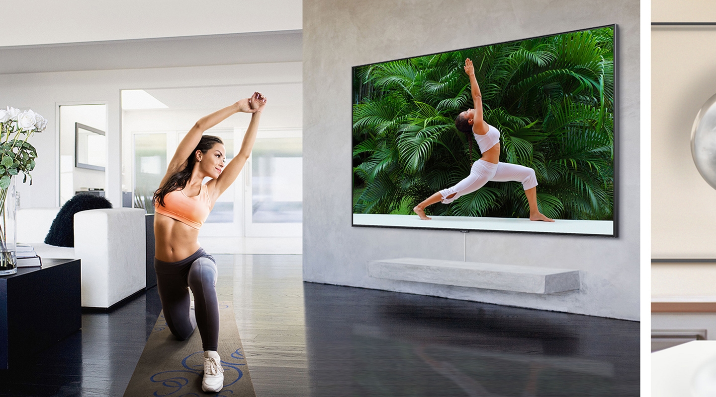 하얀색 심플한 인테리어를 한 거실에서 스포츠웨어를 착용한 여성이 초대형 TV 화면에서 요가 트레이닝 프로그램을 보면서 요가 자세를 따라갑니다.