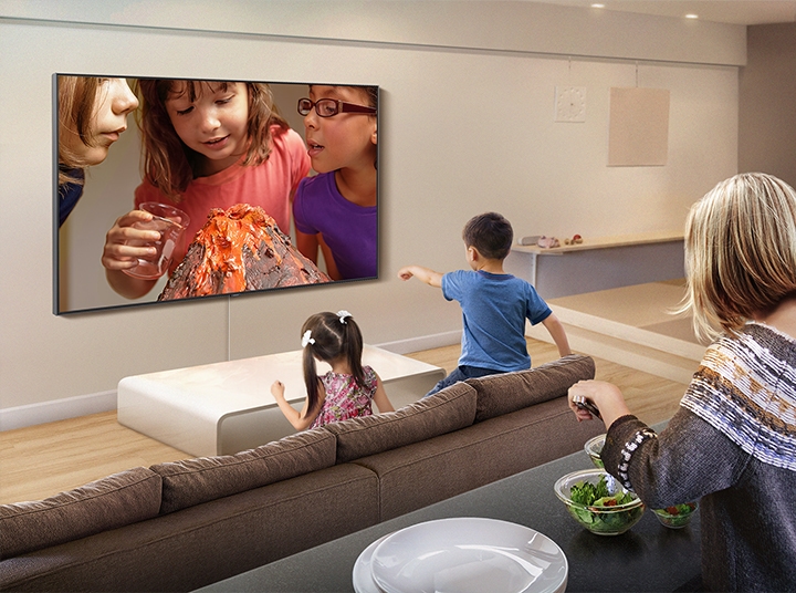 아이들은 거실에서 아이들의 과학 실험 영상을 보고 있고, 그 뒤에는 어머니가 섬 식탁에서 샐러드를 대접하고 있습니다. 실험 영상은 삼성 스마트 TV의 유튜브 앱을 통해 스트리밍되고 있습니다.
