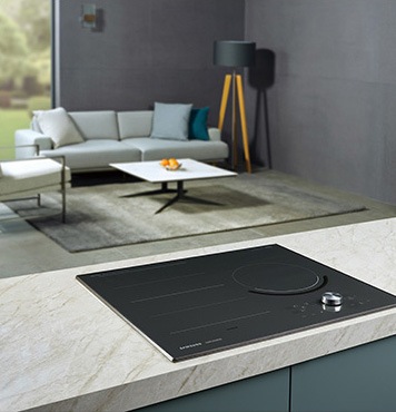 대리석으로 된 조리대 위에 전기레인지가 빌트인 방식으로 설치되어 있으며 뒤로 보이는 거실에는 밝은색의 소파와 사각테이블이 놓여 있습니다.