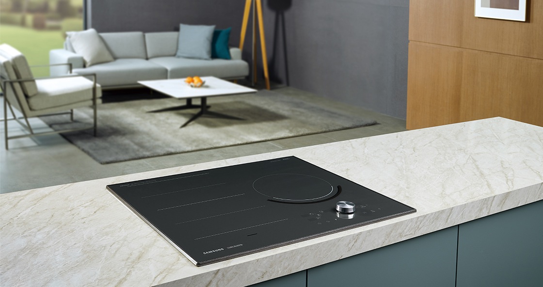 대리석으로 된 조리대 위에 전기레인지가 빌트인 방식으로 설치되어 있으며 뒤로 보이는 거실에는 밝은색의 소파와 사각테이블이 놓여 있습니다.