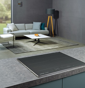 대리석으로 된 조리대 위에 전기레인지가 빌트인 방식으로 설치되어 있으며 뒤로 보이는 거실에는 밝은색의 소파와 사각테이블이 놓여 있습니다.