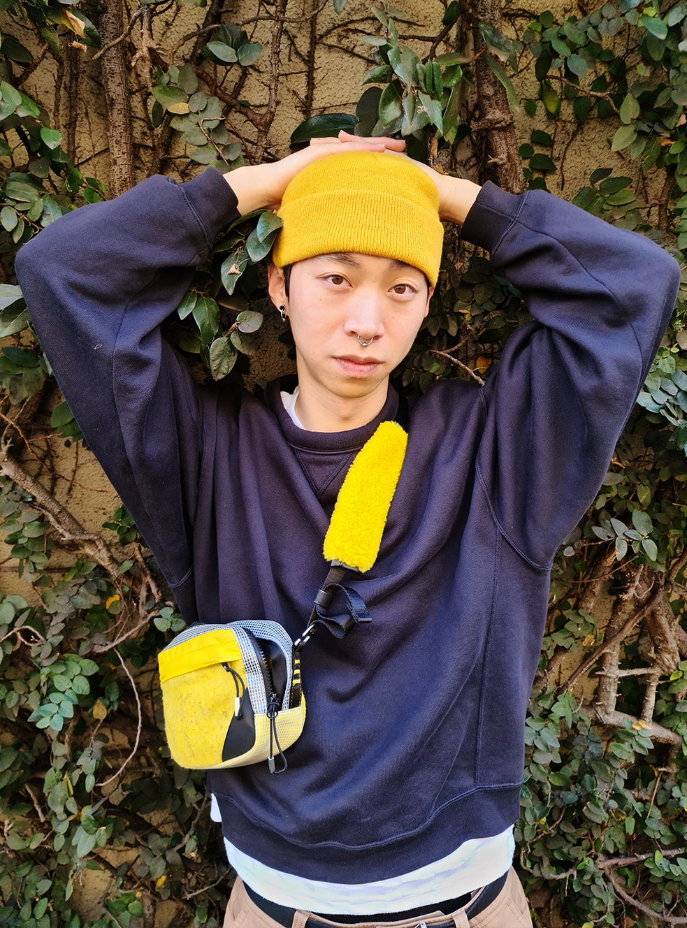 A man is posing in the background of plants wearing a navy blue clothe with a yellow hat and a bag as a point.