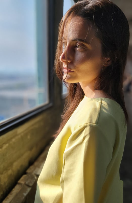 A photo image of a woman in a yellow top looking through a window with sunlight coming in.