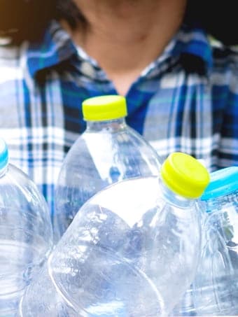 A woman wearing a blue checkered shirt is holding a box of several empty plastic bottles.
