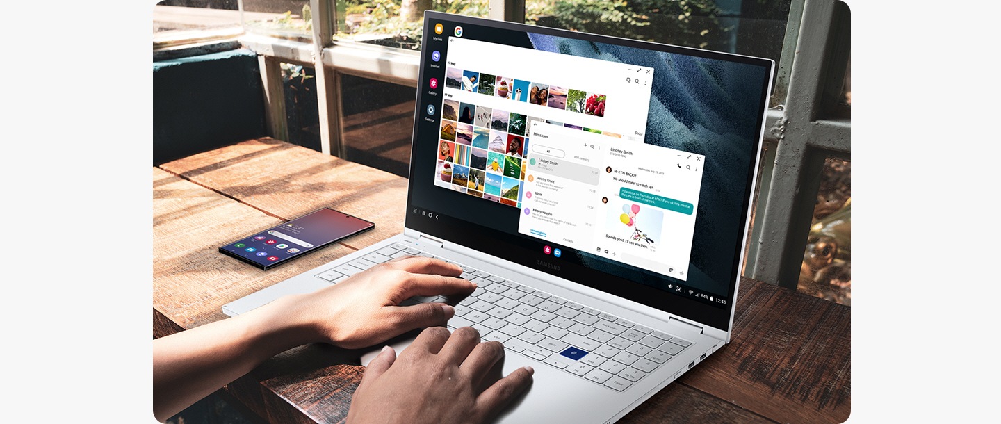 A Samsung laptop is being used. Next to the laptop is a Galaxy smartphone device showing the home screen. With Dex, the user is using the laptop screen to see the gallery while also sending text messages.