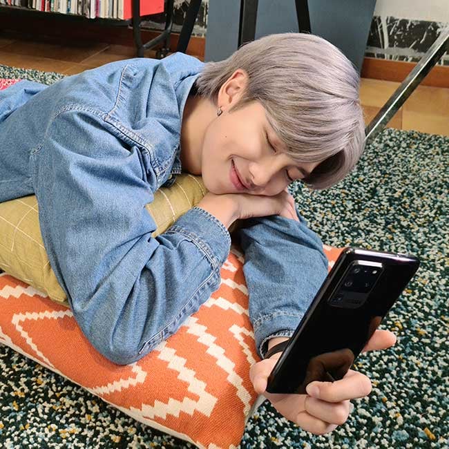 RM from BTS lays on his front on a pile of cushions on the ground, his eyes are closed and he is holding out a Galaxy S20 in front of him