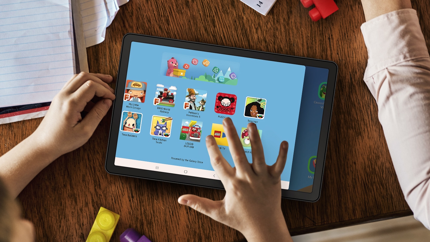 A tablet on a table surrounded by papers and LEGO blocks. A child’s hands are nearby and their right hand is touching the tablet screen. Onscreen is the LEGO App UI, showing different kinds of entertainment your child can experience.