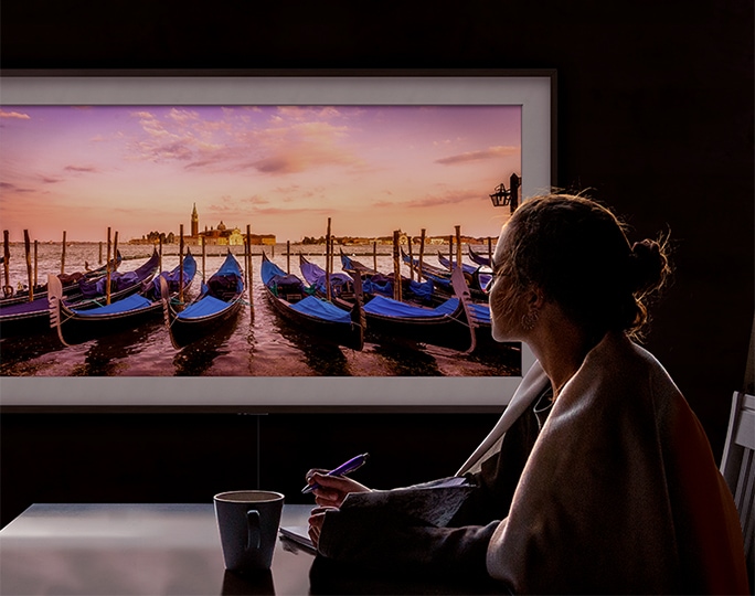 노트와 펜을 손에 들고 의자에 앉은 여성이 벽에 설치된 삼성 The Frame의 아트모드로 Serge Ramelli의 작품인 Sunset Over Gondolas를 감상합니다.
