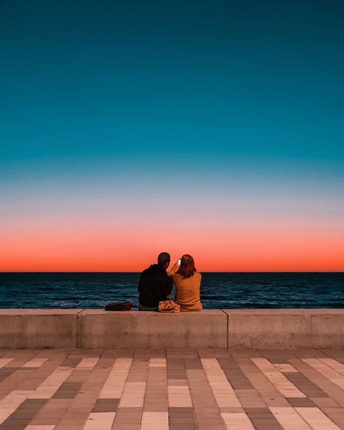 남녀가 콘크리트 난간 위에 앉아 해 질 녘의 바다를 보고 있습니다. 푸른 하늘과 바다의 경계에는 붉은 빛으로 물들어 있습니다.