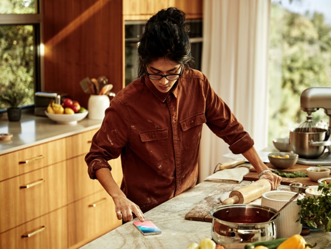 한 여성이 부엌에서 요리를 하며 온 스크린 지문 인식을 통해 스마트폰 잠금 해제를 하는 이미지입니다.  