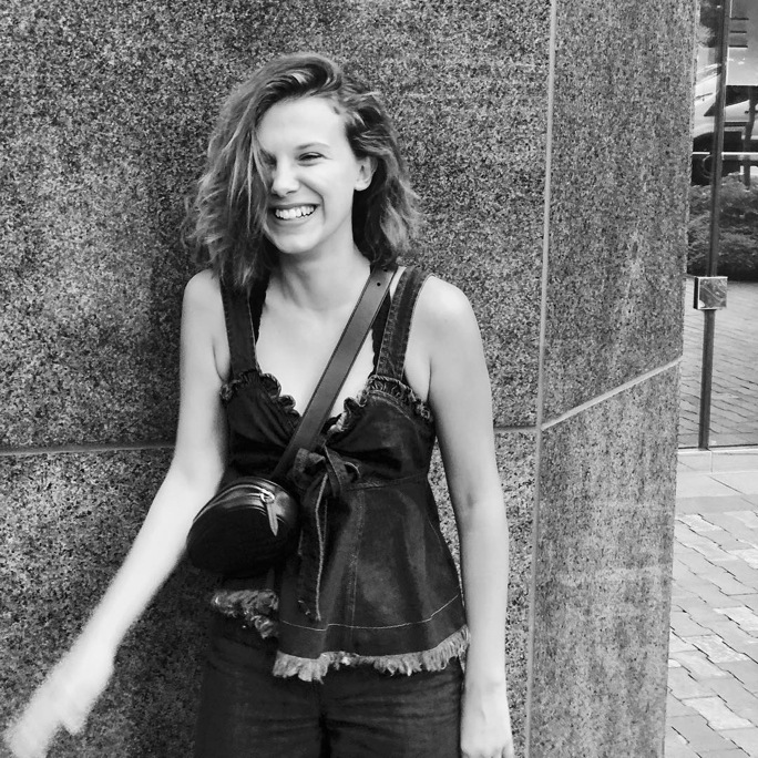 데님 탑과 청바지를 입은 밀리 바비 브라운이 길거리에서 환하게 웃으며 긍정적인 에너지를 보여주는 흑백 사진입니다.