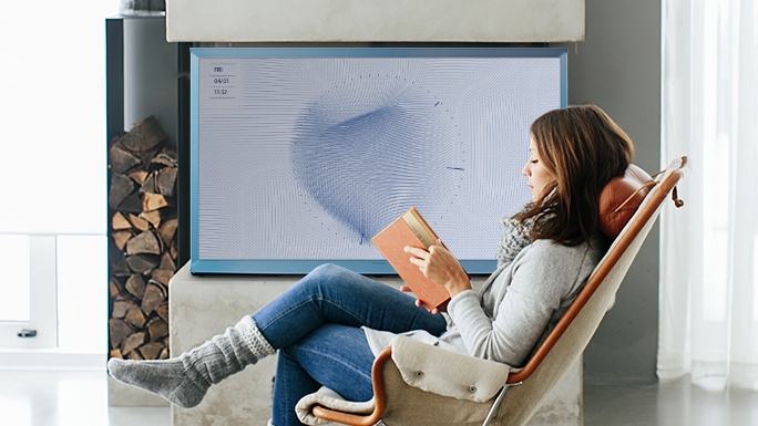 한 여성이 라운지 체어에 앉아 책을 읽고 있습니다. 여성의 뒤에는 벽난로와 The Serif 코튼 블루가 있습니다. TV 화면에는 매직스크린+으로 파란색 나뭇잎 패턴이 띄워져 있습니다.