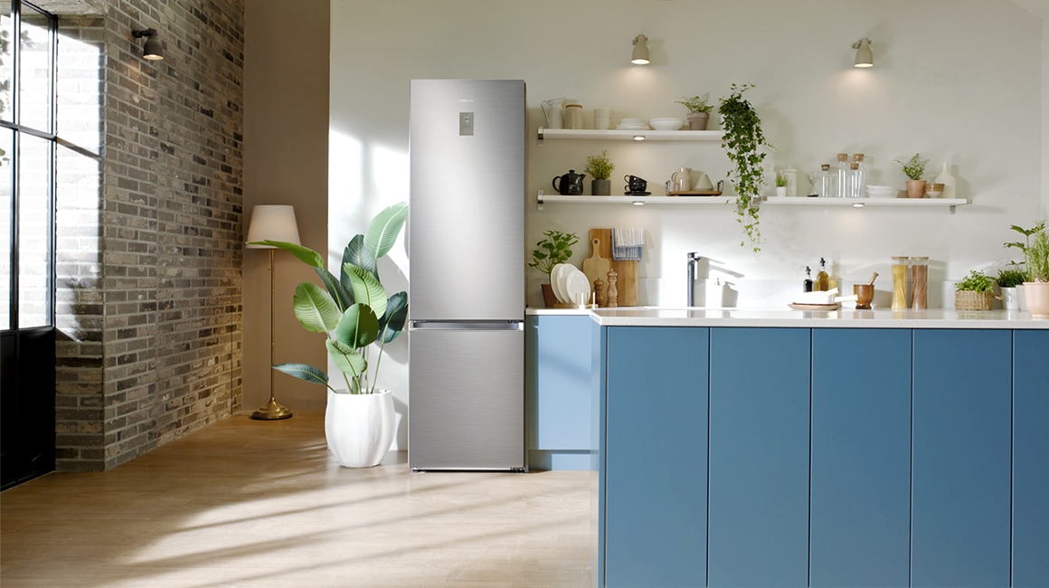 A Samsung fridge is standing in between a green plant and cadet blue sinks In the modern minimalist kitchen. And shelves stacked with some plates and decorated with small plants.