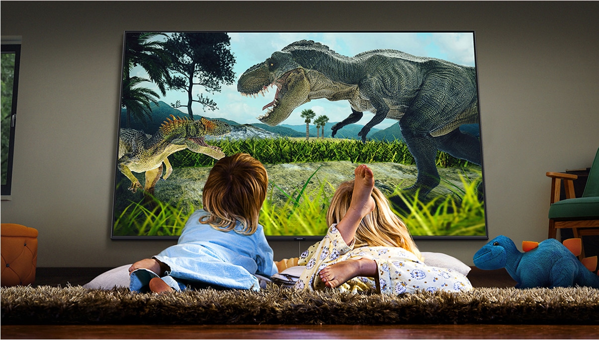 잠옷을 입고 바닥에 누워있는 두 어린이가 대형 스크린 TV로 실감나는 공룡 영화를 보고 있습니다.