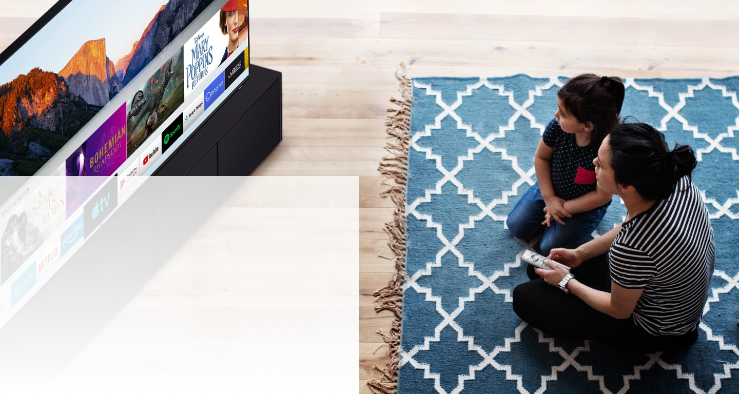 Sedute su un tappeto blu a motivi bianchi, la donna e la bambina dell’immagine stanno guardando uno smart TV di Samsung. In particolare, stanno usando le diverse app installate sul televisore.