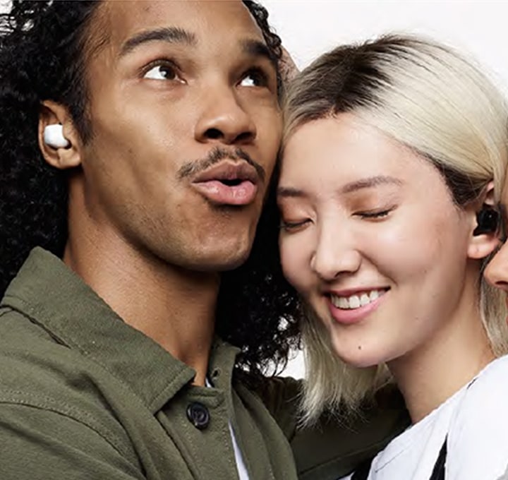 A close-up of three friends in a tight hug. A man on the left is wearing white earbuds and looks up while speaking. A woman in the middle wearing black earbuds closes her eyes and smiles. A woman on the right wearing blue earbuds rests her head on the middle woman's shoulder and smiles with her eyes closed. The Ambient sound volume GUI on the top right is turned on to the loudest setting.