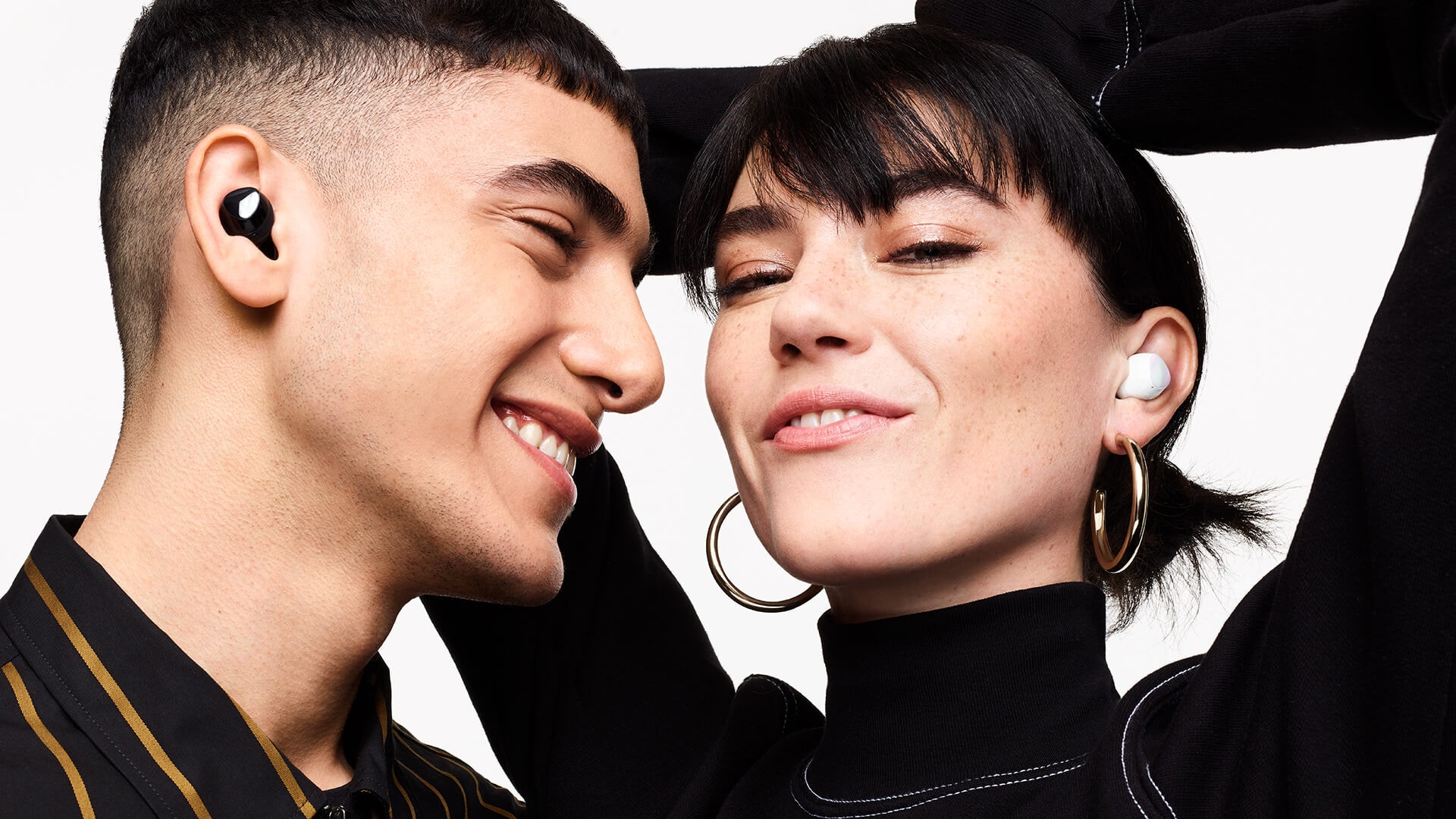 A man wearing black Galaxy Buds plus smiles as he looks a woman next to him wearing white Galaxy Buds plus. The woman smiles with her hands in the air behind her head.