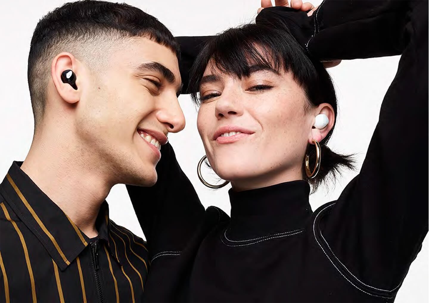 A man wearing black Galaxy Buds plus smiles as he looks a woman next to him wearing white Galaxy Buds plus. The woman smiles with her hands in the air behind her head.