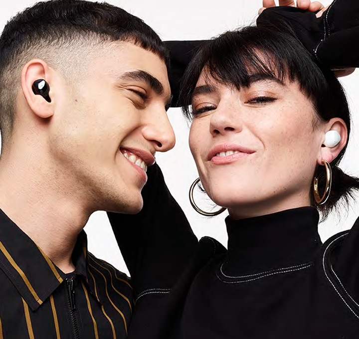 A man wearing black Galaxy Buds plus smiles as he looks a woman next to him wearing white Galaxy Buds plus. The woman smiles with her hands in the air behind her head.