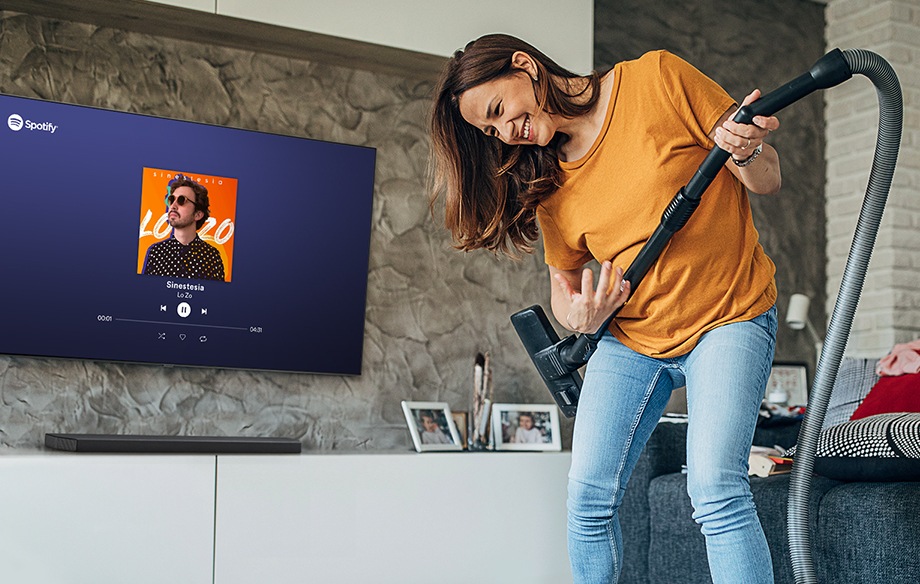 A woman uses her vacuum as a guitar while listening to Sinestesia by Lo Zo on her Samsung Smart TV.