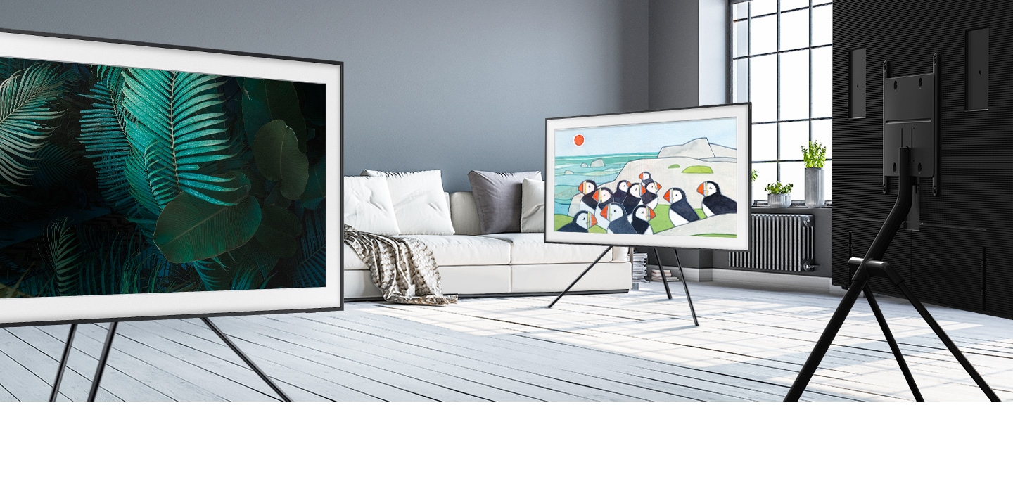 Three TVs placed on Studio Stand are in various positions in a large and stylish living room with a white sofa and window. The TV in the middle by the sofa has a painting of penguins onscreen, while the TV on the left has a display of green leaves. On the right, the third TV is facing away.