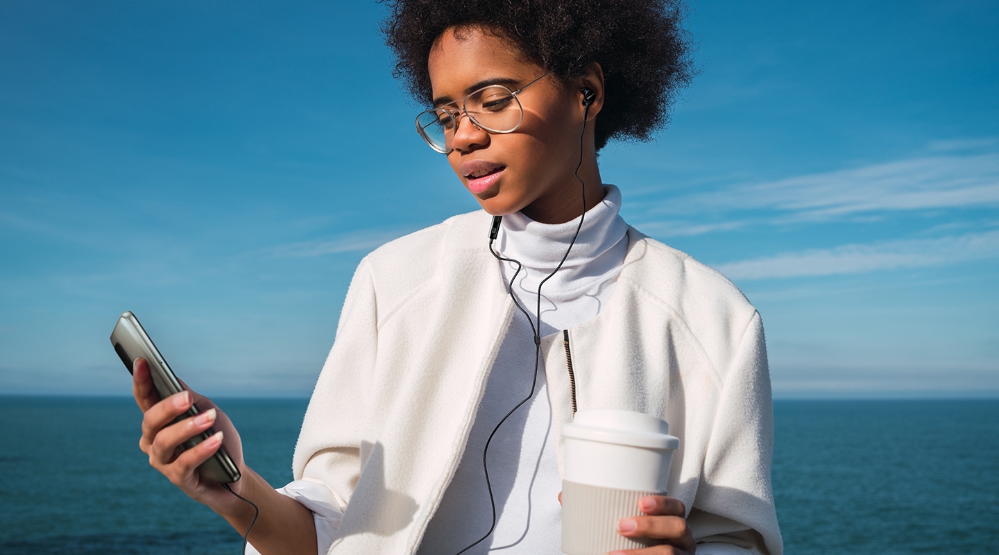 A woman is listening to music using Samsung 3.5mm earphones in the blue sea and sky background. She is holding a cup in one hand and a mobile phone connected with earphones in the other.